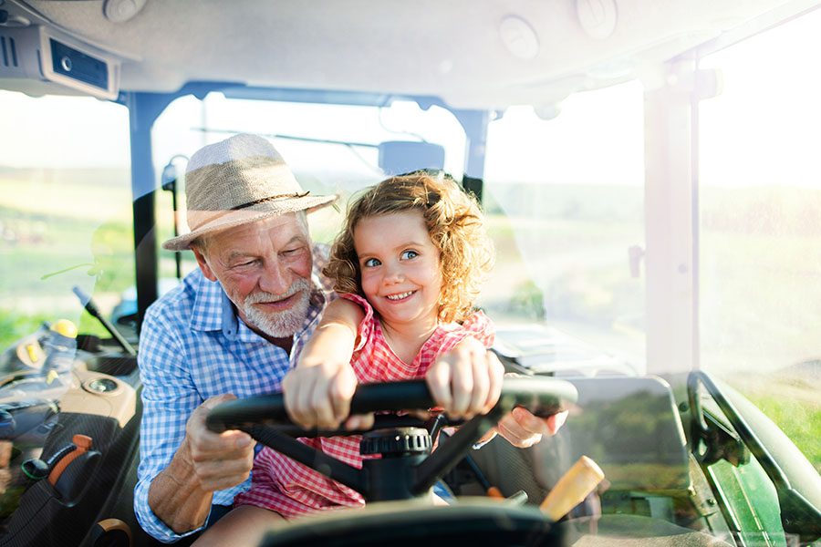 Employee Benefits - Closeup Portrait of a Cheerful Grandfather Riding in a Tractor with His Excited Granddaughter on Their Farm on a Warm Summer Day