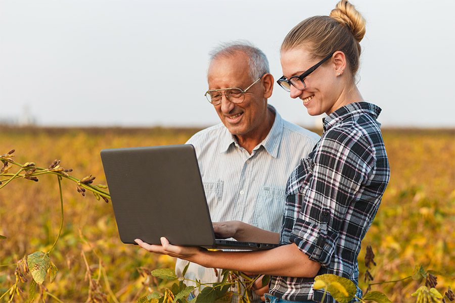 Blog - Older Man and Younger Female Smiling and Standing in a Farm Field While Looking at a Laptop on a Beautiful Day